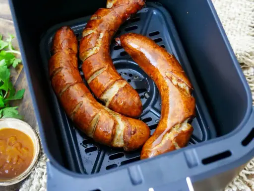 Drei frische grobe Bratwürste garen in einem Airfryer. Daneben eine Schale mit Dip und Petersilie.
