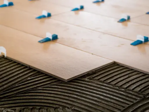 Pose de carrelage 60x60 en cours. Des carreaux en bois clair sont posés sur du mortier frais, avec des croisillons bleus et blancs pour un espacement parfait.