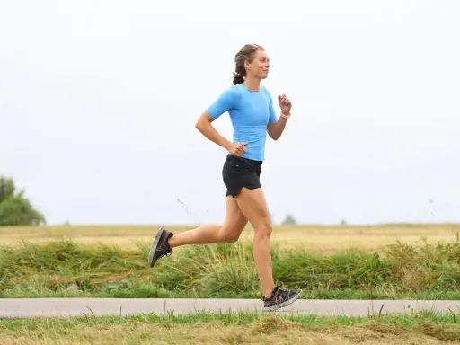 Kvinna i blå tröja och svarta shorts springer på en landsväg. Hon har en imponerande **run streak** och njuter av löprundan i det öppna landskapet.