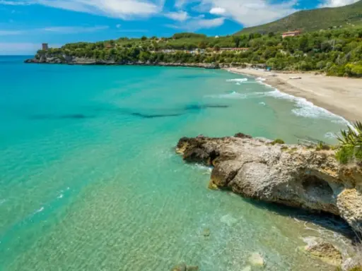 Spiaggia dorata e mare cristallino nel Cilento, con una torre antica su una scogliera e colline verdi sullo sfondo.