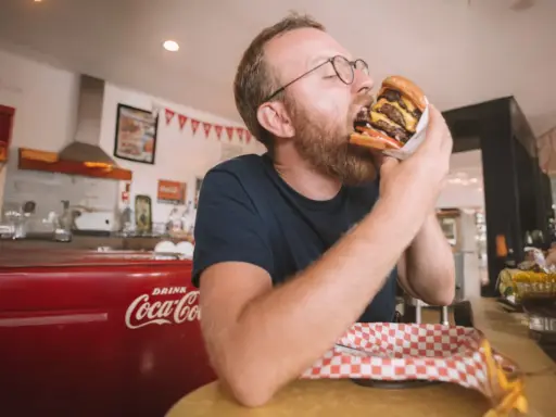 Mężczyzna z brodą i okularami zajada ogromnego burgera. W tle czerwona lodówka Coca-Coli, która może być jedną z ciekawostek o USA.