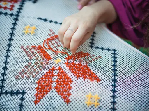 Dłoń z igłą i czerwoną nitką wyszywa wzór na białej kanwie. Widać fragmenty czerwonych i żółtych gwiazdek. Zamek otwiera się w kierunku ręki.