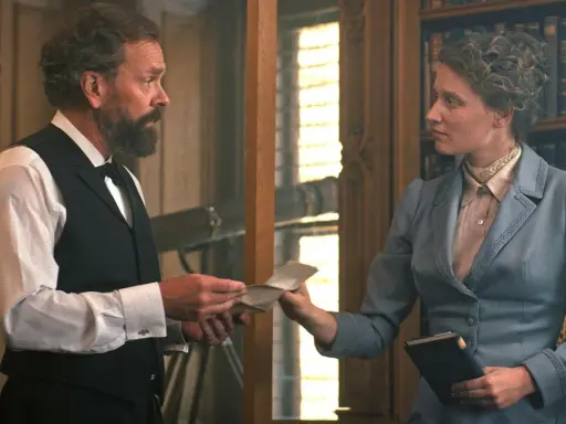 En scen från en alfred nobel film. En man med skägg ger ett brev till en kvinna i en blå dräkt framför en bokhylla.