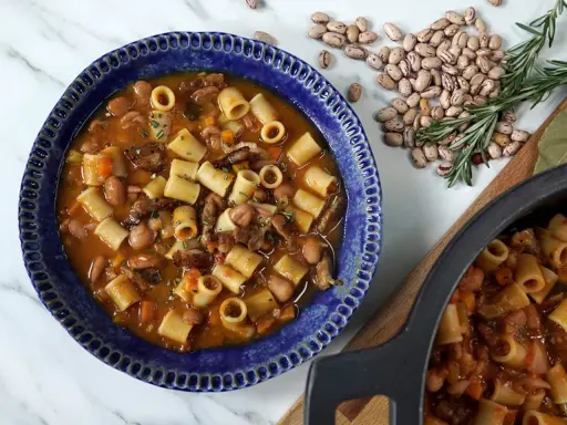 Un reconfortante plato de pasta e fagioli en un cuenco azul, con frijoles y romero esparcidos alrededor.
