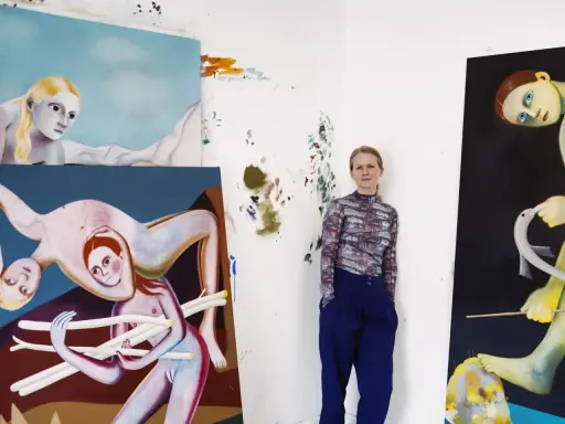 A woman stands in her studio surrounded by her surreal, figurative paintings, showcasing her as one of today's popular contemporary artists.
