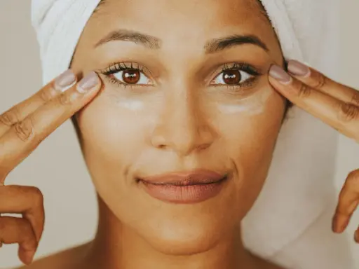 Une femme applique de la crème contour des yeux sur tout le visage, avec une serviette sur la tête.