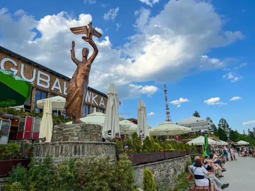 Gubałówka atrakcje: pomnik z orłem, restauracje, parasole i ludzie odpoczywający na ławkach.