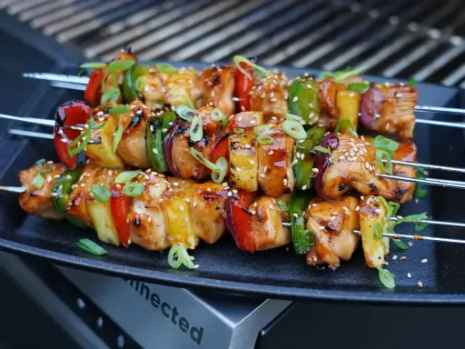 Huli Huli Chicken Spieße mit Paprika, Zwiebeln und Ananas, glasiert und mit Sesam bestreut, auf einem Grill.