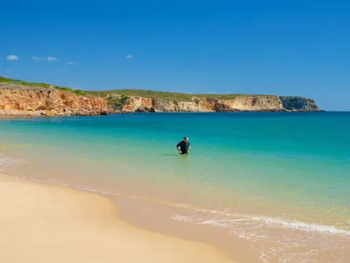 Ein Mann im Neoprenanzug steht im türkisfarbenen Wasser am Praia do Martinhal. Im Hintergrund ragen ockerfarbene Klippen auf.