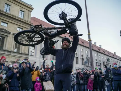 Jonas Deichmann, sichtlich erschöpft, hält sein schlammbedecktes Fahrrad über den Kopf. Eine jubelnde Menge mit Smartphones feiert seine Ankunft.