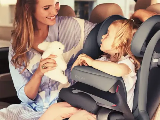 Uśmiechnięta mama z pluszowym zajączkiem przytula córkę w foteliku samochodowym Cybex Pallas 9-36 kg.