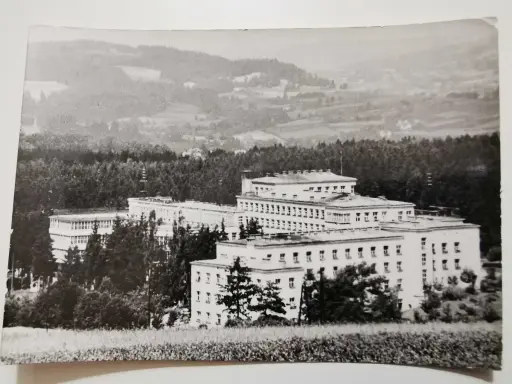 Duży budynek sanatorium w otoczeniu drzew, na tle pagórkowatego krajobrazu.