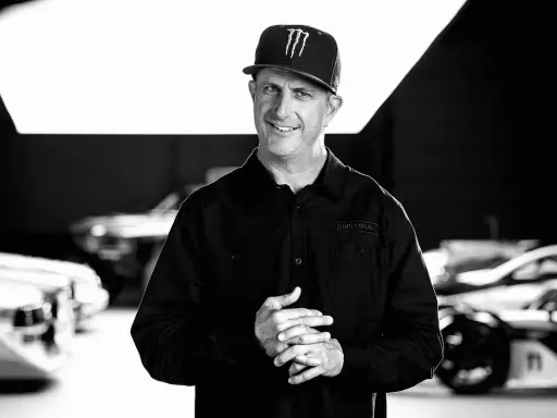 Ken Block, sonriente y con gorra, posa frente a coches de rally.