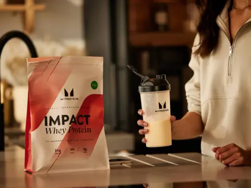 Eine Person hält einen Shaker mit einem cremigen Getränk. Daneben liegt eine Packung Myprotein Impact Whey Protein. Perfekt für den proteinshake vor dem schlafen.