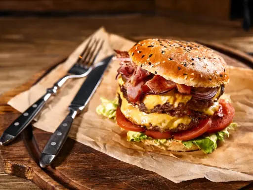 Soczysty burger z podwójną porcją mięsa, serem, bekonem i warzywami na desce z widelcem i nożem.