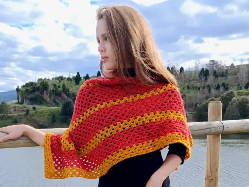 Chica con un pañuelo triangular crochet rojo y amarillo, apoyada en una barandilla de madera junto a un lago.