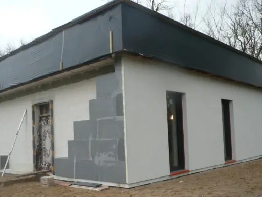 Budowa domu ze styropianu w toku. Ściany z bloczków, częściowo otynkowane, dach przykryty czarną folią.