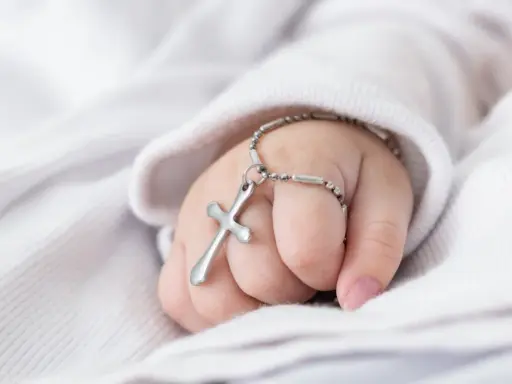 Dziecięca dłoń trzyma krzyżyk na łańcuszku, symbol tego, co jest potrzebne do chrztu.