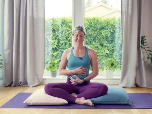 Kvinna i lila kläder sitter i meditationsställning på en yogamatta, med händerna över magen. Hon utövar yoga gravid.