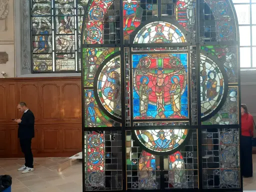 Un vitrail coloré, représentant des scènes bibliques, se dresse dans un intérieur. Un homme consulte son téléphone devant ce chef-d'œuvre.