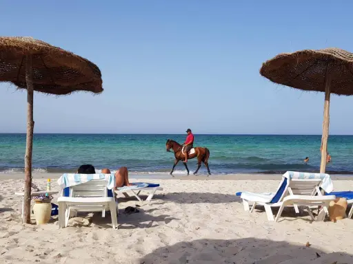 Piękna plaża na Djerbie, Tunezja. Jeździec na koniu przemierza brzeg morza, podczas gdy inni odpoczywają pod parasolami.