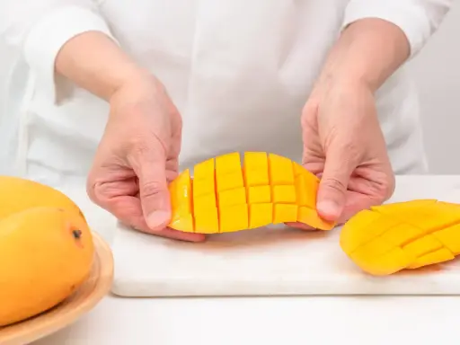 Ręce trzymają pokrojone w kostkę mango, pokazując, jak pokroić mango. Obok leżą całe owoce.