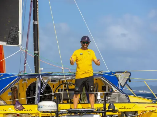 Arnaud Boissières, souriant, fait un double pouce levé sur son voilier jaune et bleu, prêt pour une nouvelle aventure en mer.