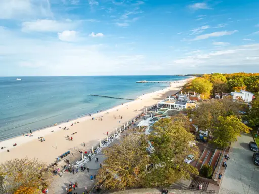 Słoneczna kołobrzeg plaża z ludźmi spacerującymi i odpoczywającymi. W tle morze i molo, a po prawej stronie drzewa w jesiennych barwach.