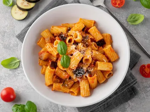 En skål med pasta alla norma, toppad med basilika och riven ost. Tomater och aubergineskivor ligger bredvid.