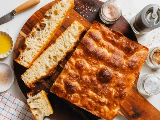 Goldbraune Focaccia mit grobem Salz, bereit zum Belegen. Daneben Olivenöl, Gewürze und ein Messer.