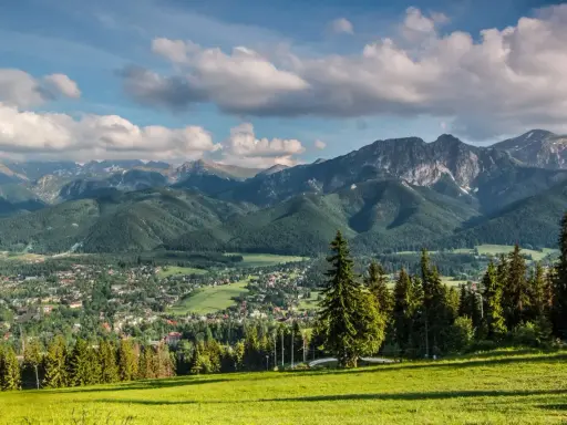 Zielona polana z widokiem na malownicze lasy w Tatrach, miasteczko i skaliste szczyty w tle.