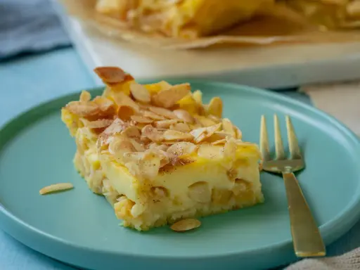 Ein Stück Apfel Schmand Kuchen mit Mandelblättchen bestreut, auf einem türkisfarbenen Teller, daneben eine goldene Gabel.