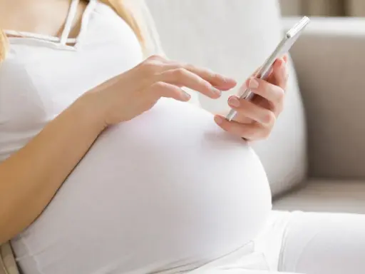 Kobieta w ciąży, w białej bluzce i kardiganie, siedzi na kanapie i korzysta z telefonu. Może sprawdzać zwolnienie lekarskie w ciąży.