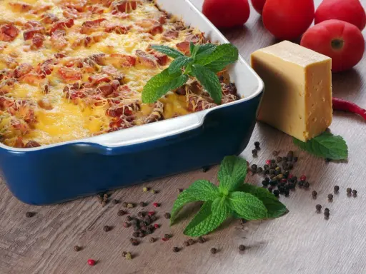 Pyszna zapiekanka makaronowa z kiełbasą, zapieczona pod złotym serem. Obok kawałek sera, pomidory i przyprawy.