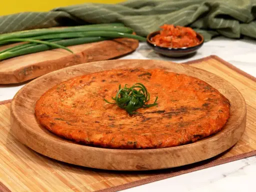 Ein goldbrauner, herzhafter Kimchi-Pfannkuchen, garniert mit Frühlingszwiebeln, liegt auf einem Holzteller. Daneben eine Schale mit Kimchi und grüne Frühlingszwiebeln.