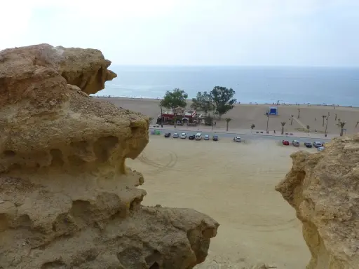 Rocas erosionadas en primer plano, con la playa de Bolnuevo, el mar y un paseo marítimo con palmeras al fondo.