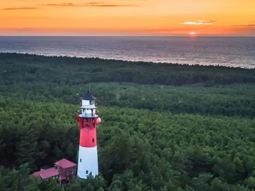 Stylowa latarnia morska w lesie, z widokiem na morze i zachodzące słońce.