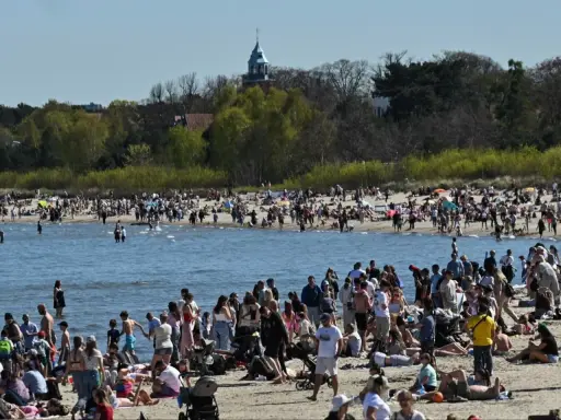 Tłumy ludzi na plaży, woda i zielone drzewa w tle. Widać wieżę kościelną. Ceny nad morzem pewnie wysokie, ale widok piękny.