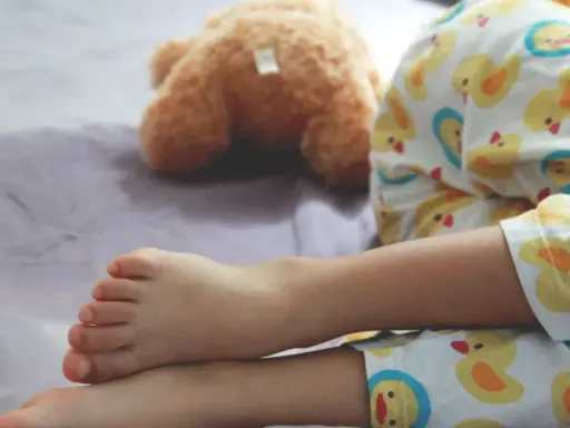 Nocne wpadki zdarzają się nawet 5-latkom. Dziecko w piżamce z kaczuszkami śpi obok pluszowego misia, a na pościeli widać mokrą plamę.