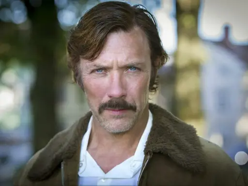 Porträtt av en svensk manlig skådespelare från 2000-talet med mustasch och blå ögon, klädd i en brun jacka med fuskpäls.