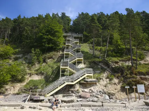 Długa, drewniana klatka schodowa prowadzi na szczyt kawczej góry. Na plaży siedzi para z psem.