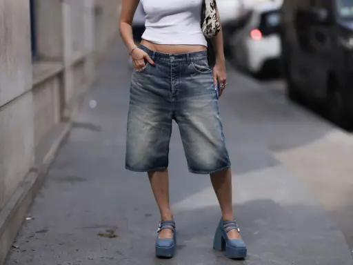 A person in denim bermuda shorts and a white crop top sports chunky blue platform Mary Janes, a nod to early 2000s shoe trends.