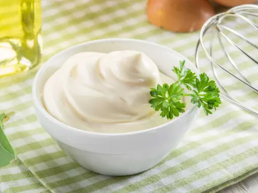 Un bol de mayonnaise maison onctueuse, garni de persil frais. Des œufs et un fouet à proximité suggèrent la préparation.