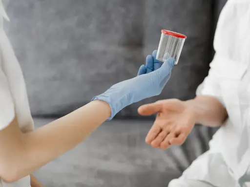 Ręka w niebieskiej rękawiczce podaje pojemnik na mocz, sugerując badanie, jak wypłukać cukier z moczu.
