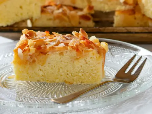 Ein Stück saftiger **Mandelkuchen vom Blech mit gemahlenen Mandeln** und Zucker bestreut, auf einem Glas-Teller mit einer Gabel.