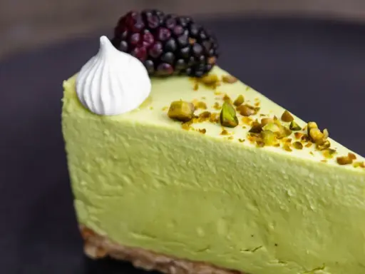 Pyszny sernik pistacjowy na zimno, udekorowany pianką, jeżyną i posypany pistacjami.