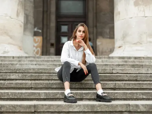 Eleganckie mokasyny to idealne buty do szarych spodni. Kobieta w białej koszuli i czarnych jeansach siedzi na schodach.