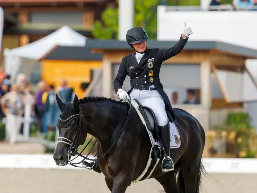 Reiterin gibt Daumen hoch nach erfolgreichem Grand Prix Reiten. Dunkles Pferd mit Turnieroutfit und Helm.
