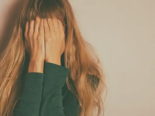 Kobieta z długimi blond włosami zakrywa twarz dłońmi na tle jasnej ściany.