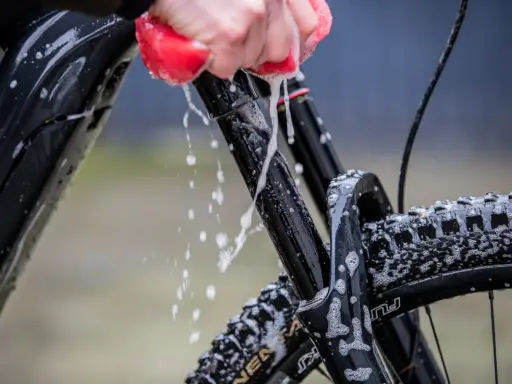 Mit Hausmitteln das Fahrrad sauber machen: Eine Hand mit rotem Schwamm reinigt den Rahmen und die Federgabel, Schaum und Wasser spritzen.